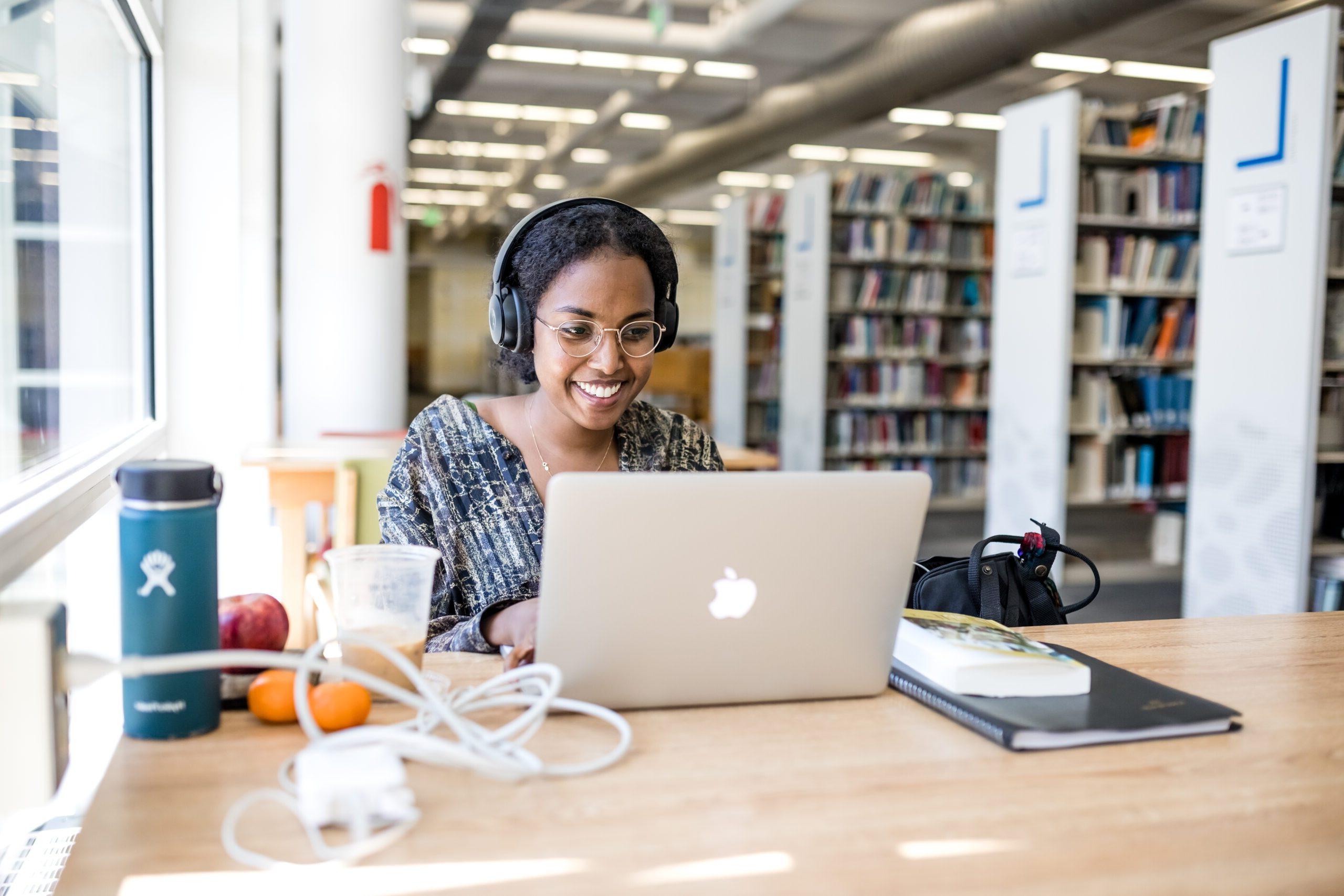 一个学生戴着耳机坐在图书馆的电脑前，面带微笑.